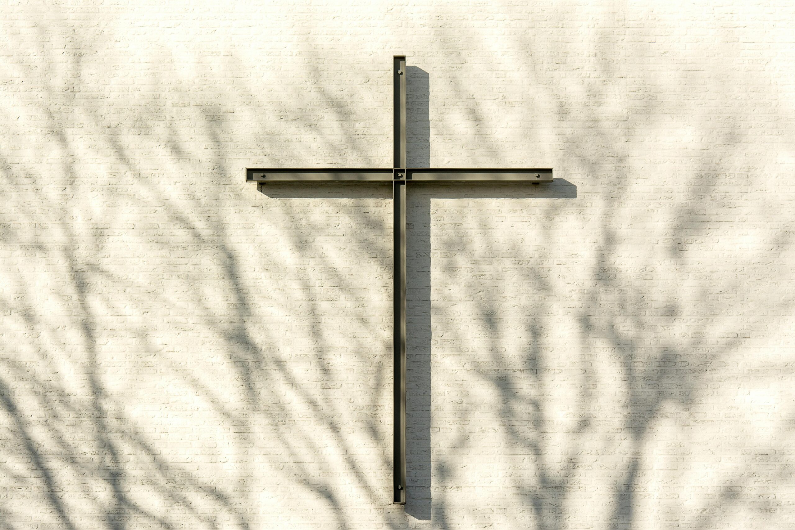 Metal cross casting shadow on a white brick wall with tree silhouettes.